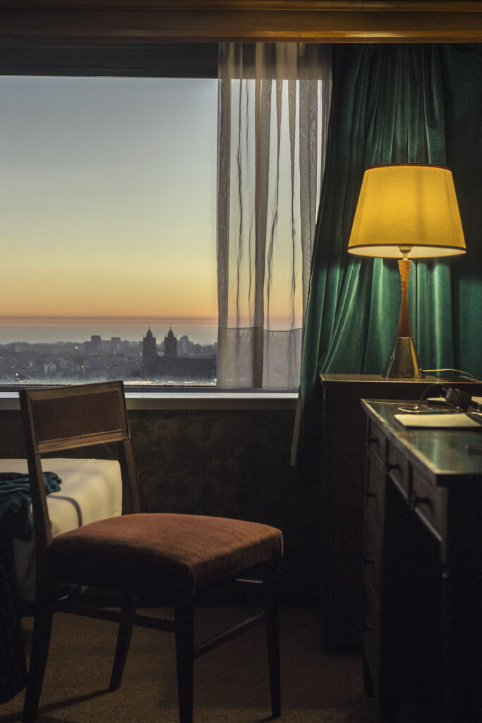 desk chair and hotel bed in front of a window with a view of a big city at sunset